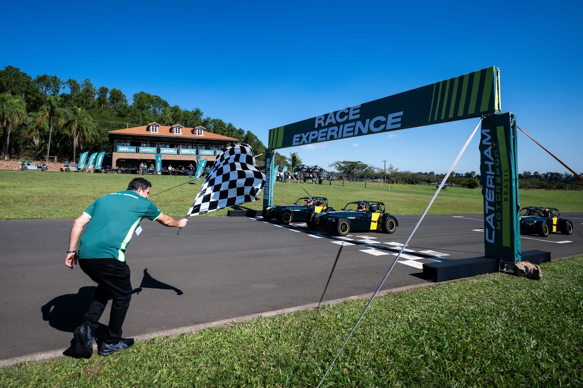 Bandeira quadriculada abaixo do pórtico Race Experience, Caterhams cruzando a linha de chegada